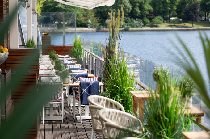 Deck 4 Terrace on the Inner Alster with comfortable chairs, striped cushions and lush greenery. Sunny, peaceful atmosphere with views of the water and trees.