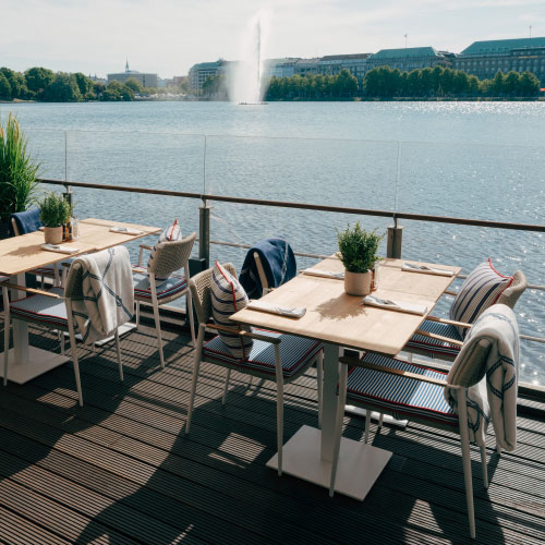 Deck 4 Terrasse an der Binnenalster mit Holztischen und -stühlen, die mit Topfpflanzen geschmückt sind. In der Ferne sprüht unter klarem Himmel ein Springbrunnen.