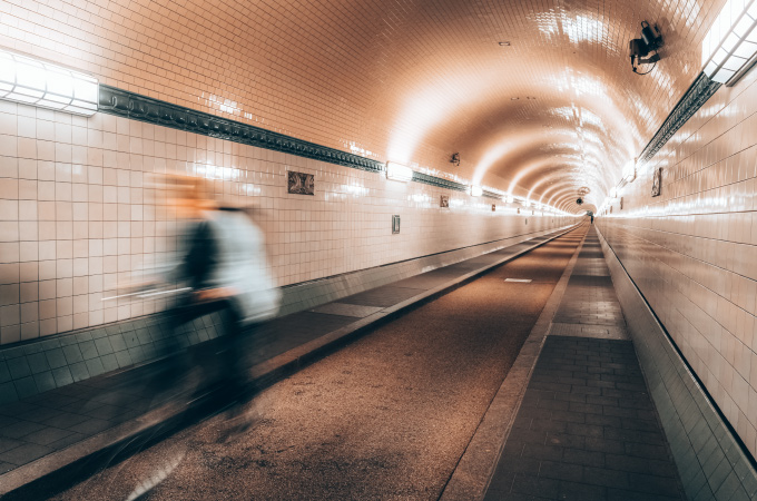 A cyclist in motion blurs past in a long, tiled underground tunnel with bright, even lighting.